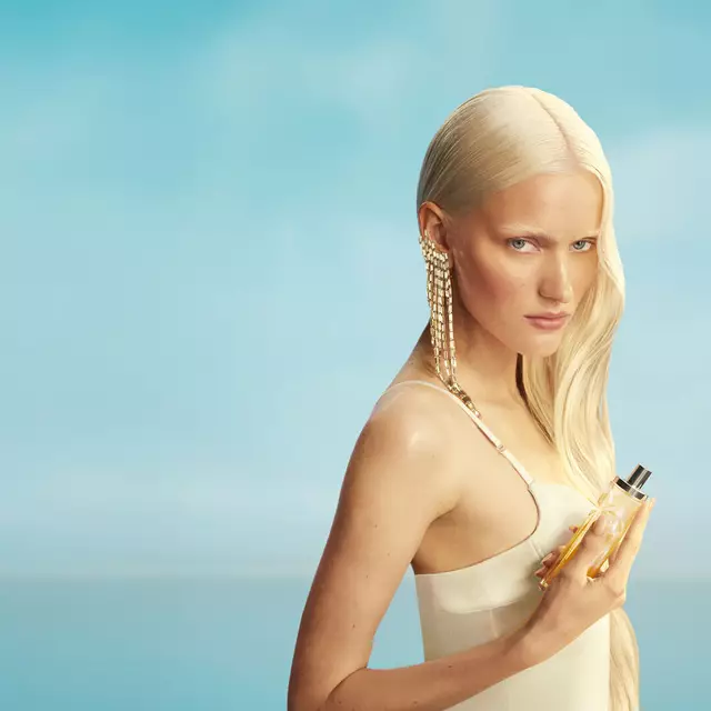 Nieuwe Mugler Starlicious les eaux gourmandes campagnebanner met een model met lang blond haar en blauwe ogen, dat een lichtgele jurk draagt en de Pineapple Musk-parfumflacon in de hand houdt tegen een achtergrond met een zachtblauwe lucht. De geuren zijn heerlijk, verzamelbaar en kunnen in laagjes worden gebruikt.