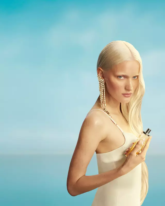 Nieuwe Mugler Starlicious les eaux gourmandes campagnebanner met een model met lang blond haar en blauwe ogen, dat een lichtgele jurk draagt en de Pineapple Musk-parfumflacon in de hand houdt tegen een achtergrond met een zachtblauwe lucht. De geuren zijn heerlijk, verzamelbaar en kunnen in laagjes worden gebruikt.